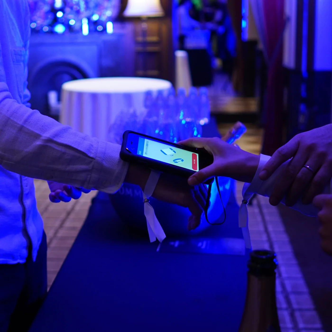 A staff member holds a ruggedised Famoco handheld against a guest's NFC wristband at a bar counter, under blue event lighting.